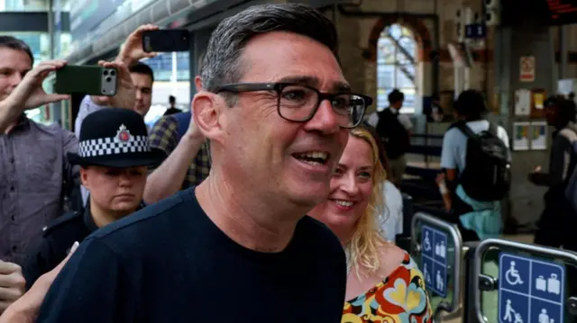 Andy Burnham, ladeado por policiais. Há multidões de pessoas tirando fotos atrás dele. Ele usa óculos e uma camisa escura.