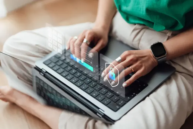 Seorang sedang menggunakan laptop di atas pahanya, dan muncul berbagai macam hologram terkait dengan keamanan digital.