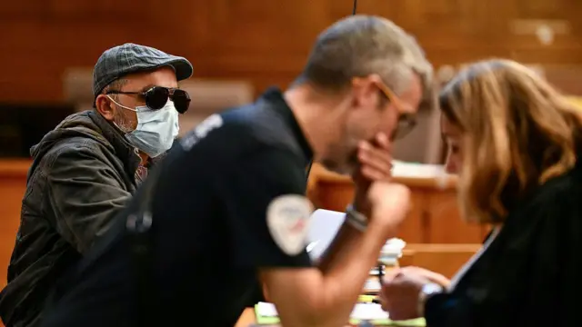 Siyah güneş gözlüğü ve maske ile yüzünü mahkeme salonunda gizleyen bir adam görüyoruz