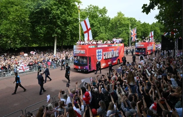 Автобусы со спортсменками едут по огороженному участку улицы, с двух сторон от ограждений стоят приветствующие их люди