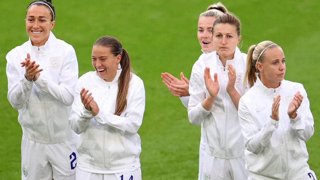 Jogadoras de futebol usando uniforme branco aplaudem enquanto sorriem