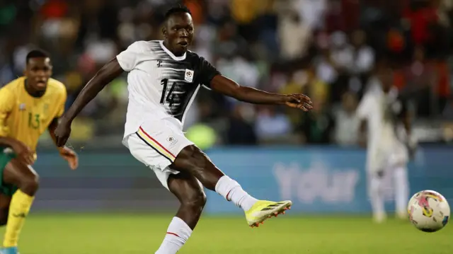L'attaquant ougandais Denis Omedi tire du pied droit en direction du but, le ballon se trouvant dans le coin inférieur droit de l'image.