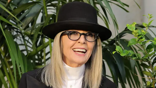 Diane Keaton luce un sombrero y un traje negro elegante y sonríe mirando a un costado rodeada de plantas ornamentales.