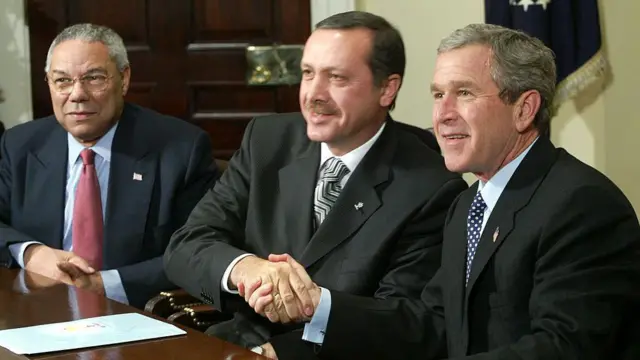 Dönemin ABD Başkanı George W. Bush (Sağda, dönemin ABD Dışişleri Bakanı Colin Powell (Solda) ve dönemin AKP Genel Başkanı Recep Tayyip Erdoğan (Ortada) 10 Aralık 2002'de, Beyaz Saray'da görüşüyor. Üç siyasetçi de oturuyor. Erdoğan ve Bush el sıkışıyor.