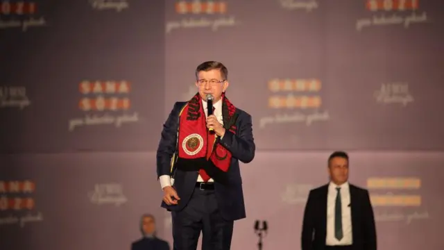 Davutoğlu kırmızı bir atkıyla bir miting sahesinde elinde mikrofonlar ayakta, resmi giyimli. 