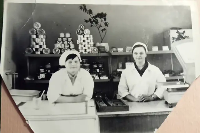 Чорно-біле фото, зроблене всередині магазину або кулінарного відділу. На передньому плані – прилавок з механічними вагами. За прилавком стоять двоє жінок у білих халатах і головних уборах. На задньому фоні – дерев’яні полиці, заповнені товарами: піраміди консервних банок, скляні банки з продуктами, кілька коробок