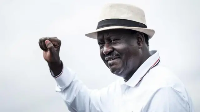 Raila Odinga wearing a hat and white shirt raises his fist to supporters