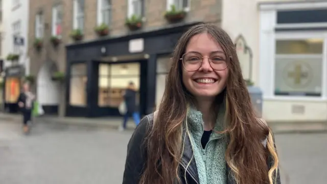 Jasmine Clement está de pie en la calle, sonriendo a la cámara. Tiene el pelo largo y castaño y gafas. Lleva una chaqueta de cuero negra sobre un forro polar verde con cremallera.