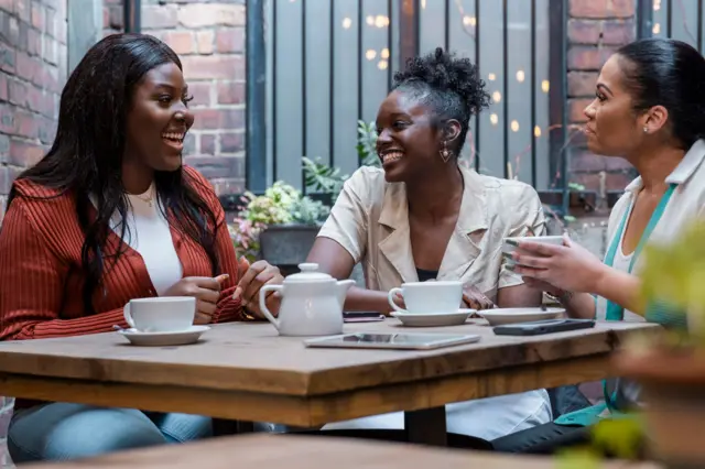 Plan moyen de trois jeunes femmes prenant un café ensemble sur une terrasse dans le nord-est de l'Angleterre. Elles rient et discutent.