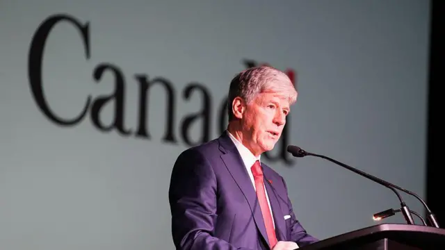 Тім Годжсон у костюмі стоїть перед мікрофоном і трибуною, позаду видно напис Canada 