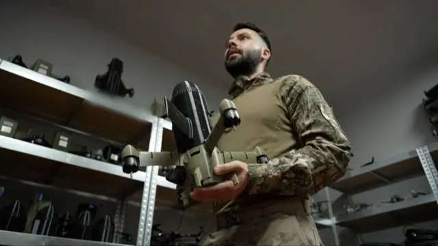 Mężczyzna w mundurze wojskowym trzyma zielono-czarny plastikowy dron przechwytujący, który ma kształt małej rakiety z czterema wystającymi ramionami. W tle na półkach widać kolejne części drona.