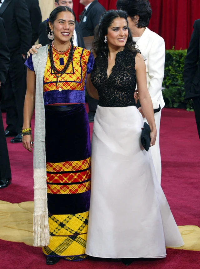 Lila Downs y Salma Hayek posan sonrientes frente a las cámaras en la alfombra roja. Downs lleva un vestido tradicional de México de colores amarillo, azul, negro y rojo. Hayek lleva un vestido largo de colores blanco y negro