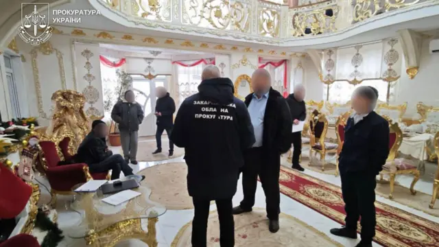 У великій залі з багатим біло‑золотим інтер’єром стоять кілька чоловіків, зайняті розмовою та переглядом документів. У центрі кадру видно співробітника в куртці з написом "Закарпатська обласна прокуратура". Дехто сидить у червоних кріслах поруч зі скляним столиком, на якому лежать папери. Приміщення прикрашене позолоченими елементами, декоративними поручнями на другому рівні та килимами з візерунками, що створює атмосферу розкоші.