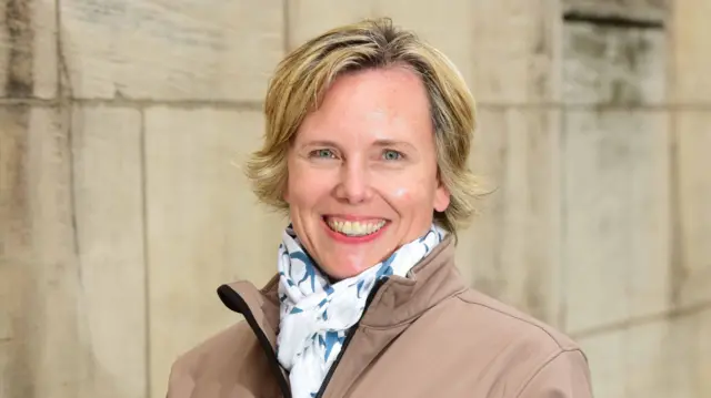 Andrea Selley tiene el pelo rubio hasta las orejas. Sonríe y lleva una bufanda blanca y azul y una chaqueta marrón claro. Está de pie frente a un muro de piedra.