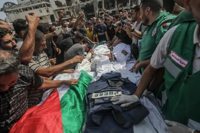 Una multitud rodea los cuerpos de los reporteros muertos en el ataque israelí, que están cubiertos por la bandera palestina y un chaleco antibalas en el que se lee "Prensa". Al fondo se ven edificios en ruinas. 