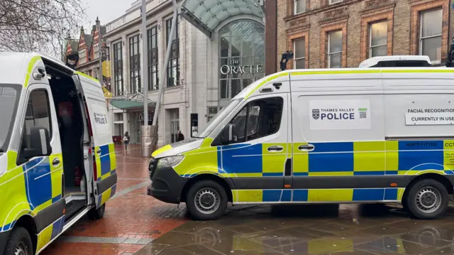 Two police vans, each with two facial recognition cameras mounted on the roof are parked in the pedestrianised Broad Street outside the Oracle shopping centre entrance in Reading. The sky is grey and the pavement is wet.