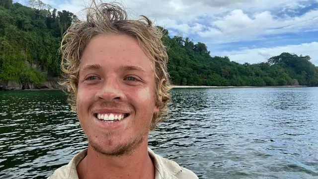 Tom Robinson sonriendo a la cámara. Detrás se ve el mar y la vegetación en la cosa de una isla del Pacífic.