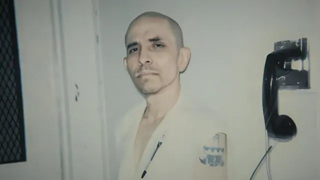 César Fierro en la cárcel con ropa de recluso. Mira hacia la cámara. Está de pie al lado de un teléfono.
