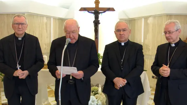 Quatre ecclésiastiques se tiennent debout, l'un d'eux parle dans un micro, un autel blanc avec des fleurs et un crucifix se trouve derrière eux.