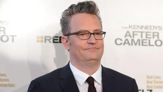 Matthew Perry wear glasses and a dark suit. E pose for picture for one red carpet event.