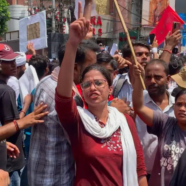 Varuni Poorva, une jeune femme qui aspire à devenir professeure, lève la main lors d'une manifestation. Elle porte un haut rouge brodé de fleurs blanches et une écharpe blanche sur les épaules. Elle est entourée d'autres manifestants.