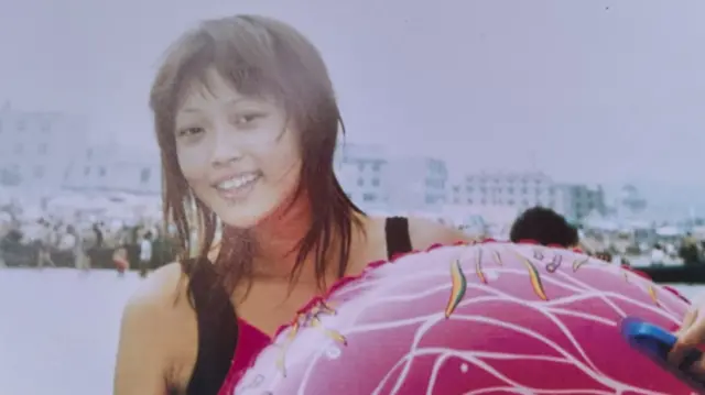 Umwigeme afise umushatsi ushika ku rutugu, yambaye agahuzu ko kwogana, ahagaze afise umupira wo kwifashisha koga mu mazi menshi, inyuma yiwe hari plage.