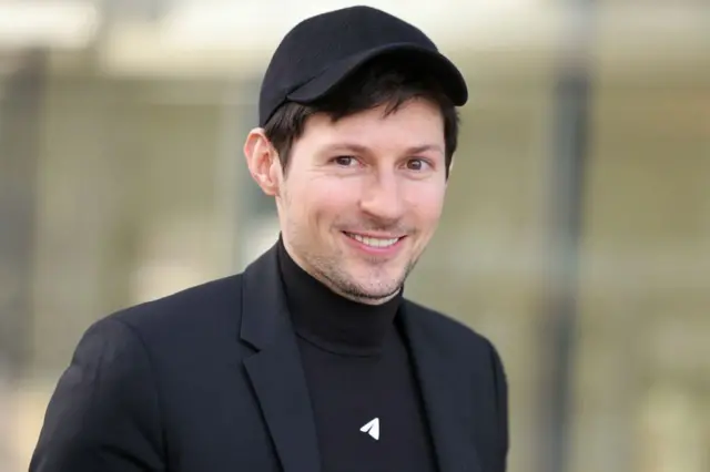 Pavel Durov sonríe y mira a la cámara. Viste un traje negro con una gorra y una camisa de cuello tortuga del mismo color.