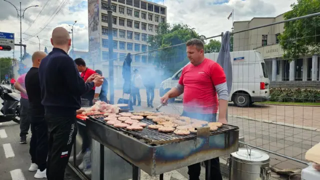 Muškarac u crvenoj majici okreće pljeskavice na roštilju na trgu