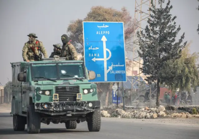 Soldiers in uniform on a military vehicle pass a sign on a road with directions to different Syrian cities