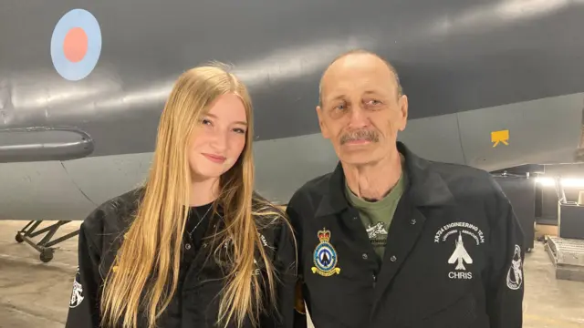 Grace ma długie, proste blond włosy i jest ubrana jest w czarny kombinezon. Uśmiecha się do obiektywu. Stoi obok swojego dziadka Chrisa, który ma siwe wąsy i nosi zielony T-shirt oraz czarny kombinezon. Oboje stoją przed samolotem.