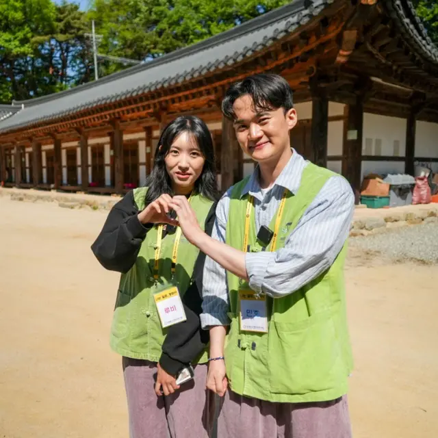 루비와 민호가 연두색 조끼와 행사 명찰을 착용한 채 마당에 서서 손을 맞잡아 하트 모양을 만들고 있다.