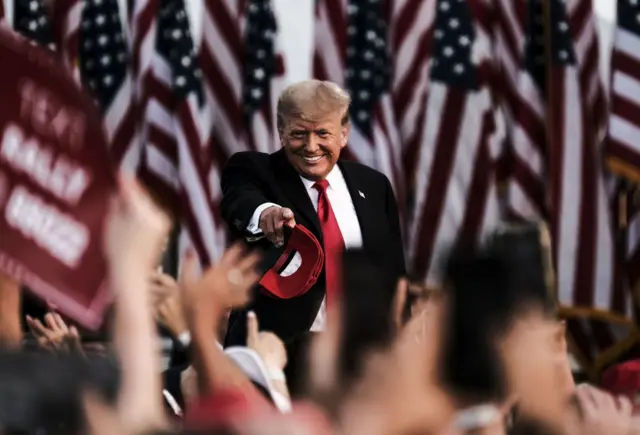 Donald Trump apontando para multidão em comício em 2021, com várias bandeiras dos EUA atrás dele