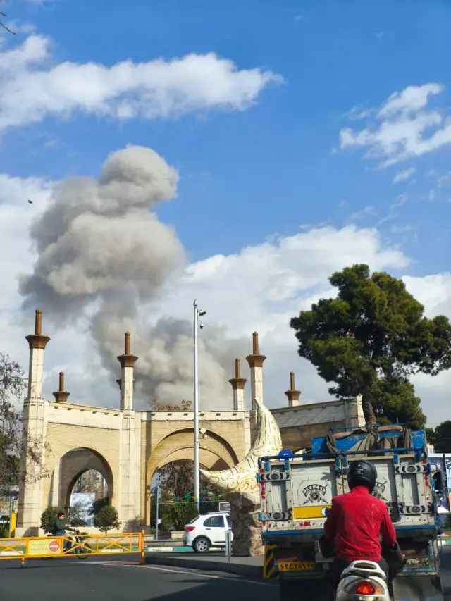 Ifoto igaragaza umwotsi w'umweru uzamuka hejuru y'umusigiti muri Irani, nyuma y'igitero cy'Amerika na Israel kuri Irani. Imbere hari umugabo uri kuri moto, hirya hari imodoka. 