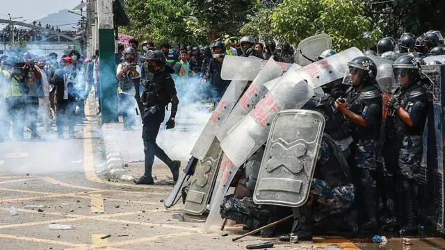 A group of police wearing helmets with visors and holding riot shields stand in the street in formation as gas or smoke billows around their legs, on 8 September 2025. None are wearing breathing apparatus or masks.