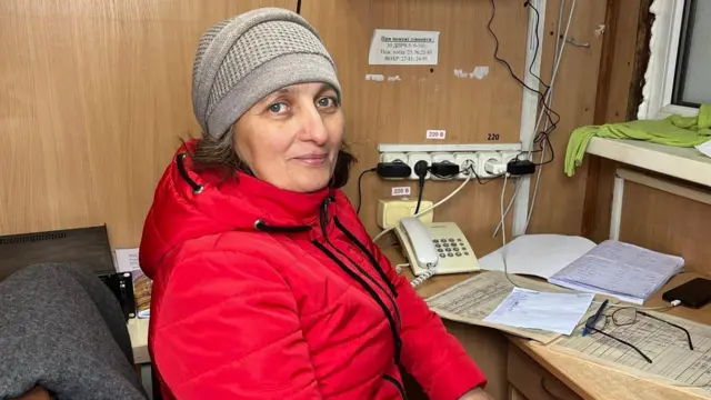 Ірина сидить у вузькій будці за столом у напівоберта. Вона вдягнена в червону куртку та сіру утеплену шапку