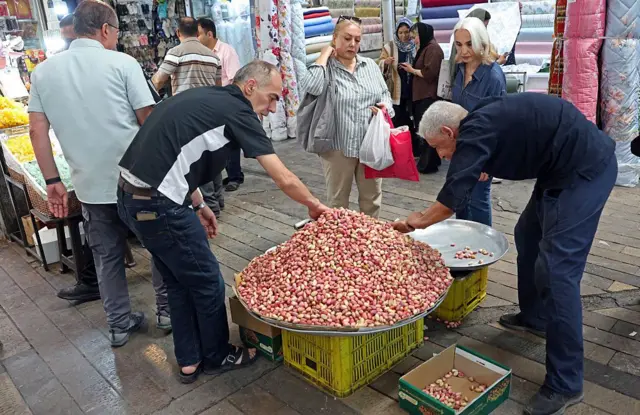 Un hombre vende granos de color rojo y blanco en la salida de un mercado. Otro, vestido de azul, toma una cantidad para comprar.