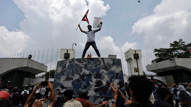 Manifestantes na porta do parlamento. Um homem está em cima de uma estrutura segurando uma bandeira