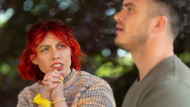 Se ve a Jessie Stride, concursante de Traidores, charlando con su fiel compañero Matthew. Jessie tiene el pelo rojo brillante, sombra de ojos amarilla y naranja, y lleva un jersey arcoíris. Al fondo hay árboles.