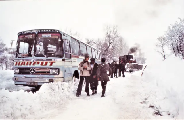 Karla kaplı yolda kalmış bir otobüs ve arkada yolu açmaya çalışan bir kar küreme makinası görülüyor. Otobüsün üzerinde Harput yazıyor. Otobüsün yanında ise bir grup insan sohbet ediyor. 