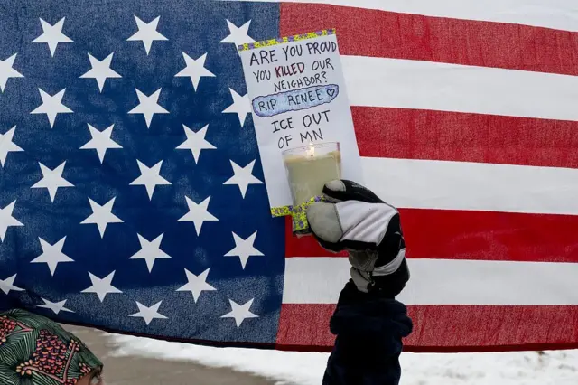 Американский флаг, перед ним рука держит свечу, к которой прикреплен лист бумаги с надписью: «Вы гордитесь тем, что убили нашу соседку?RIP Rene. ICE, прочь из Миннесоты!»