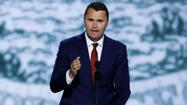 Charlie Kirk speaks at a lectern. He has cropped brown hair. He wears a dark blue suit jacket, white shirt and red tie and is mid-speech. He is standing in front of a patterned white and blue background.