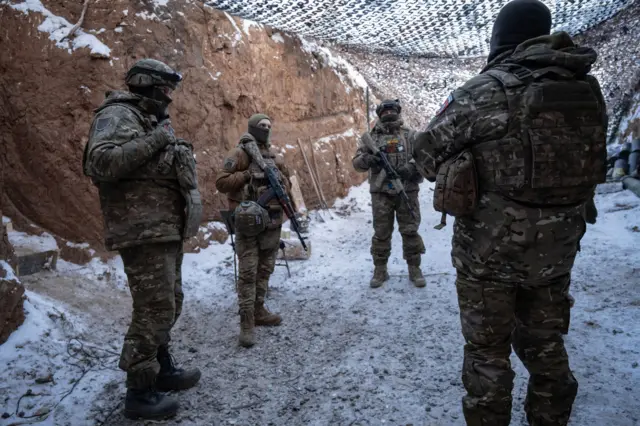 Четверо українських військових з автоматами в траншеї, накритій маскувальною сіткою 