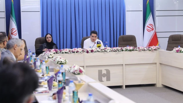 تصویر اتاق کنفرانس ساختمان سپهر بانک صادرات را نشان می‌دهد. همایون شجریان با تی‌شرت سفیدرنگ در کنار مدیر برنامه‌اش سحر فروزان که با روسری و مانتو مشکی نشسته است. در گوشه تصویر حاضران دیده می‌شوند.