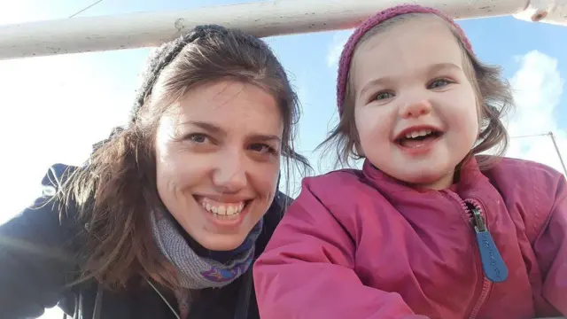 Beyaz iki korkuluğun arasında birbirine yaklaşarak gülümseyen bir kadın ve küçük bir kız, arkada mavi bir gökyüzü