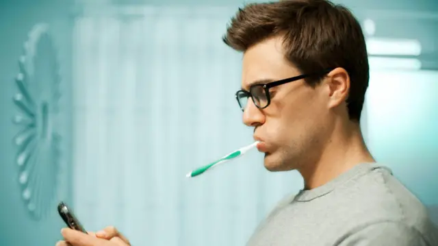 Homem de perfil no banheiro, usando óculos com uma escova de dentes na boca, enquanto consulta seu telefone celular