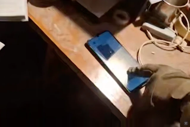 На столі лежить смартфон, на екрані якого відкрито месенджер. Поруч — рука в камуфляжній рукавичці, яка торкається телефону. На столі також є білі кабелі, подовжувач і частково видно інші предмети, зокрема металеву кружку.