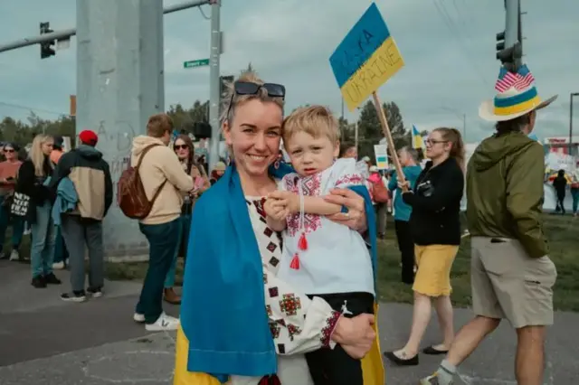 девушка в вышиванке и с ребенком на руках. На заднем плане люди с плакатами в поддержку Украины