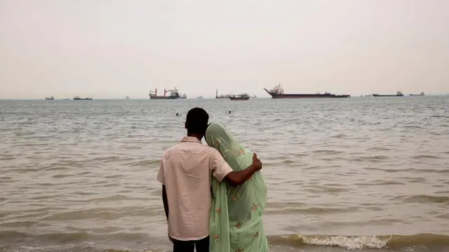Мужчина и женщина смотрят на море, а у берега стоят на якоре корабли, 22 апреля 2026 года, Бандар-Аббас, Иран.