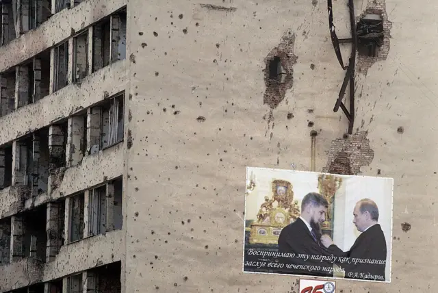 2005-жыл. Грозныйдын борборунда урушта жабыркаган үйдө Путиндин Рамзан Кадыровго сыйлык тапшырган учур тартылган плакат илинип турат