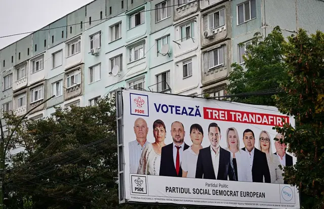 На цій фотографії зображено передвиборчий білборд партії ПСДП з написом "Голосуйте за три троянди!" напередодні парламентських виборів у місті Унгени 24 вересня 2025 року. Плакат висить на фоні дерев і житлового будинку